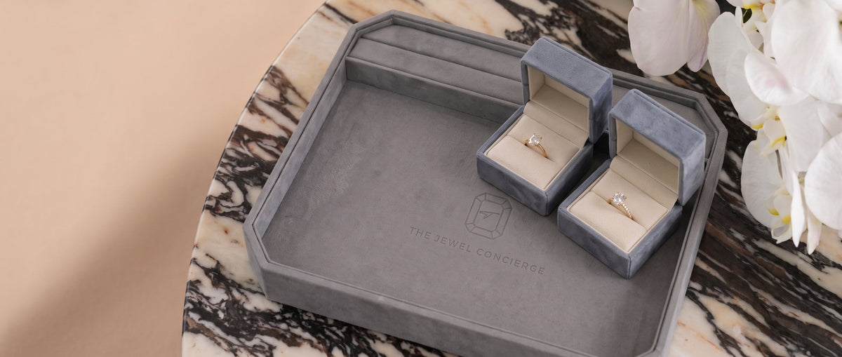 Two Lab-grown diamond engagement rings in a slate blue velvet ring box, elegantly placed on a marble table against a beige background