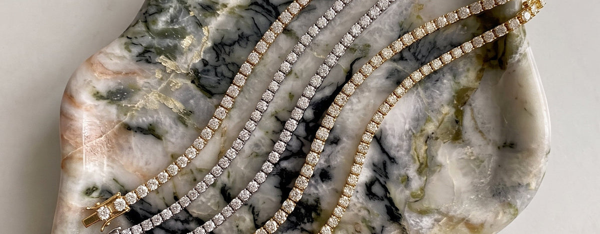 Five lab-grown diamond tennis bracelets with clear stones, arranged on a polished marbled stone tray over a light marble surface - The Jewel Concierge