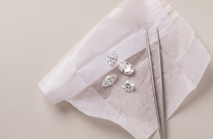 Assortment of lab-grown diamonds laid on white parchment paper along with diamond tweezers on a soft luxurious grey background - The Jewel Concierge