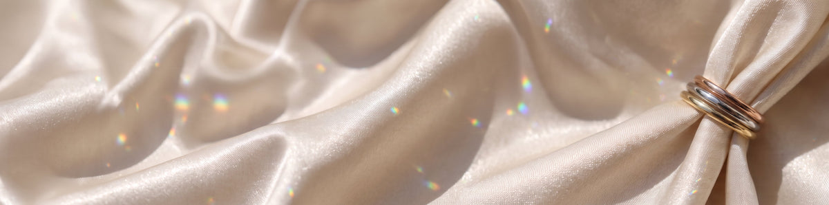 The Emily Ring - classic half rounded wedding bands, finished in yellow, white and rose gold, resting on a draped satin fabric with iridescent light flecks | The Jewel Concierge