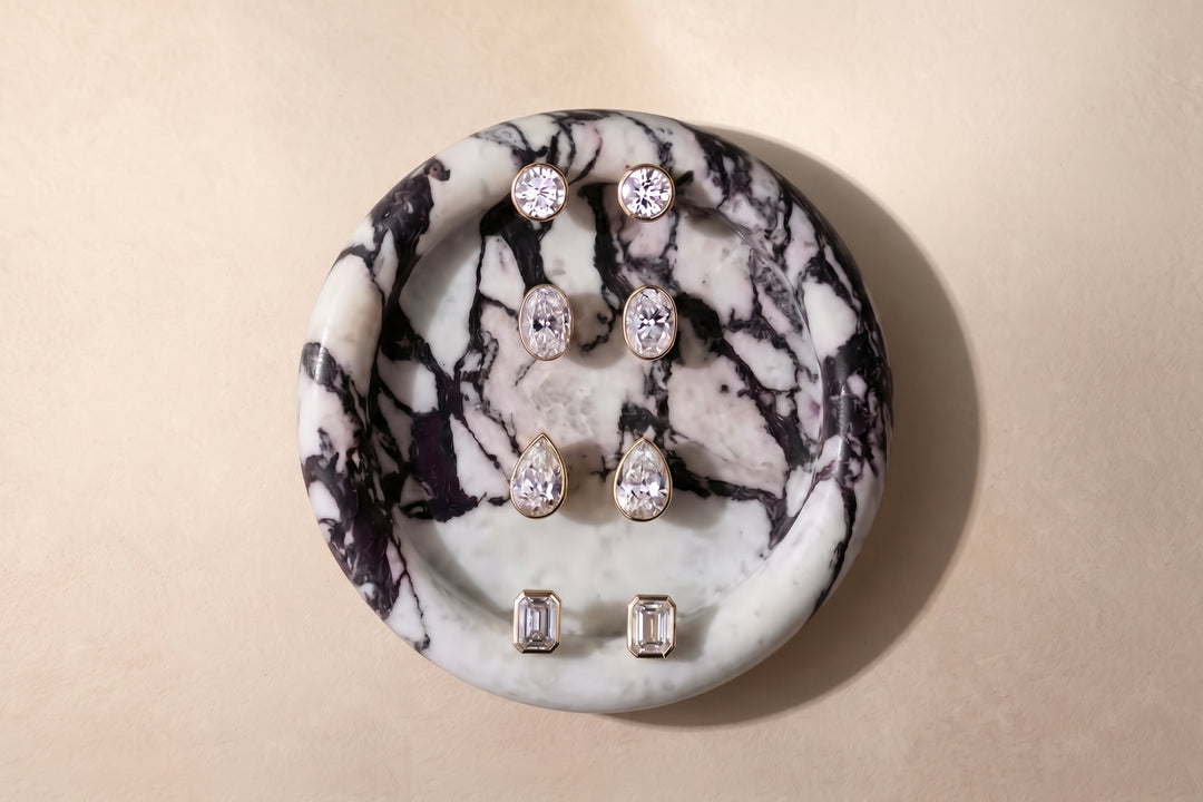 Four pairs of The Aurelia Stud Earrings displayed on a round calacatta viola marble tray against a warm beige background. The earrings feature lab-grown diamonds in a bezel setting of yellow gold, including round, pear, and emerald cuts.