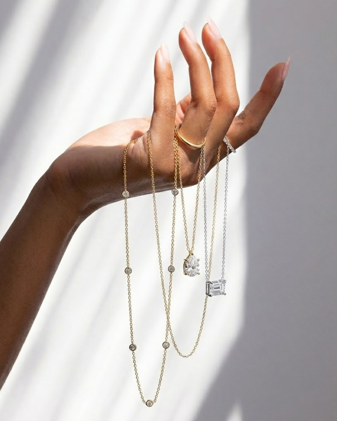 Hand wearing multiple necklaces with diamond pendants on a neutral background - The Jewel Concierge