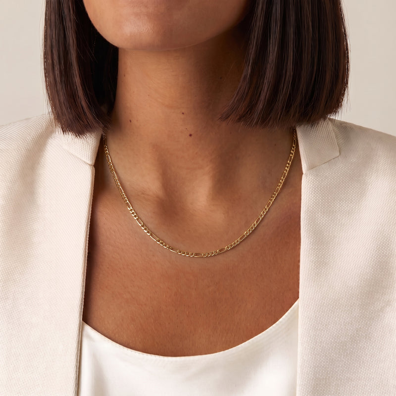 A close-up of a model wearing a delicate figaro gold chain necklace with a textured curb link design, styled with a cream-colored blazer - The Jewel Concierge