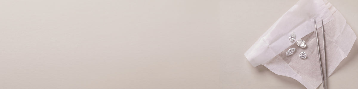 Assortment of lab-grown diamonds laid on white parchment paper along with diamond tweezers on a soft luxurious grey background - The Jewel Concierge