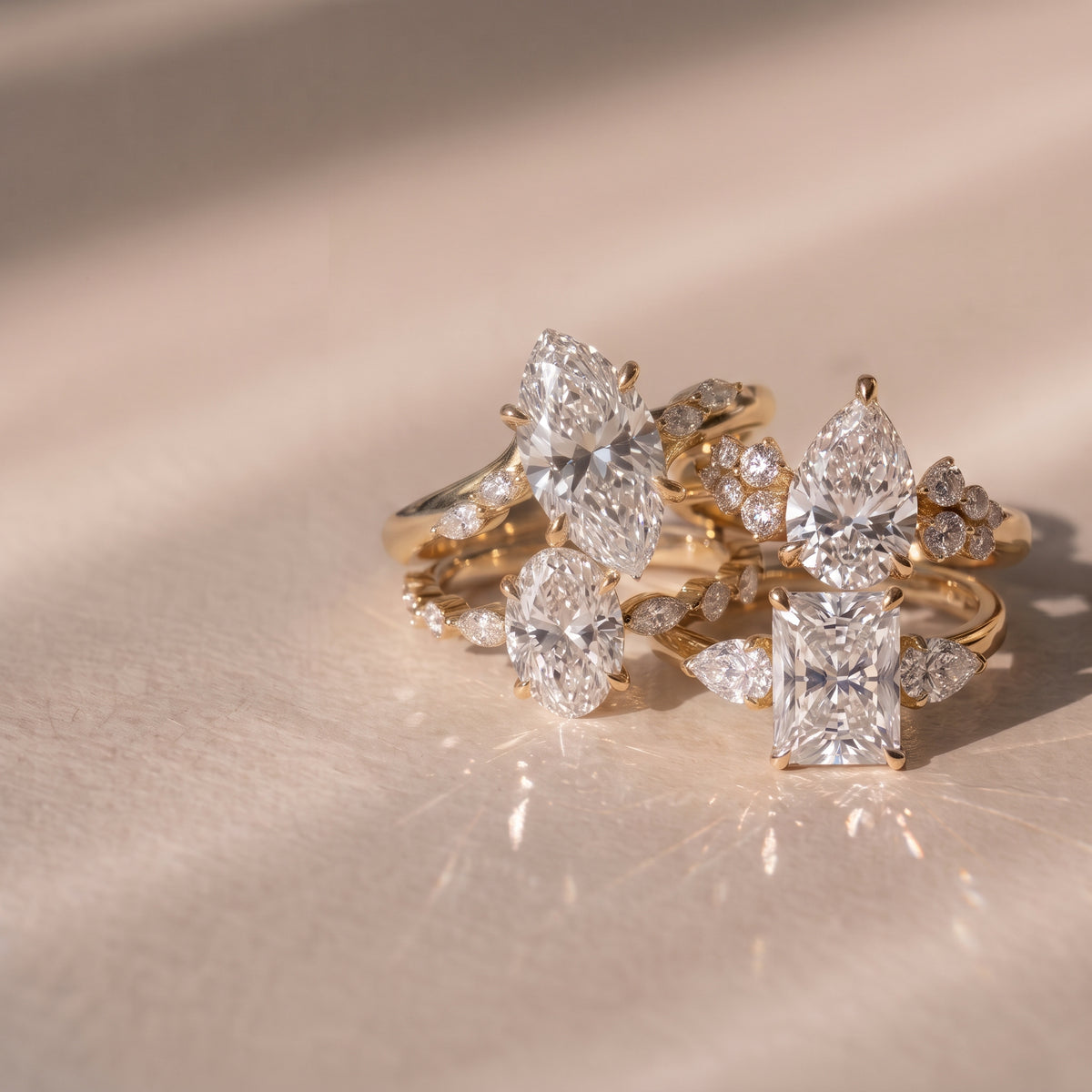 Four Lab-grown diamond engagement rings in yellow gold with different diamond shapes, elegantly resting on one another agaisnt a warm beige background | The Jewel Concierge