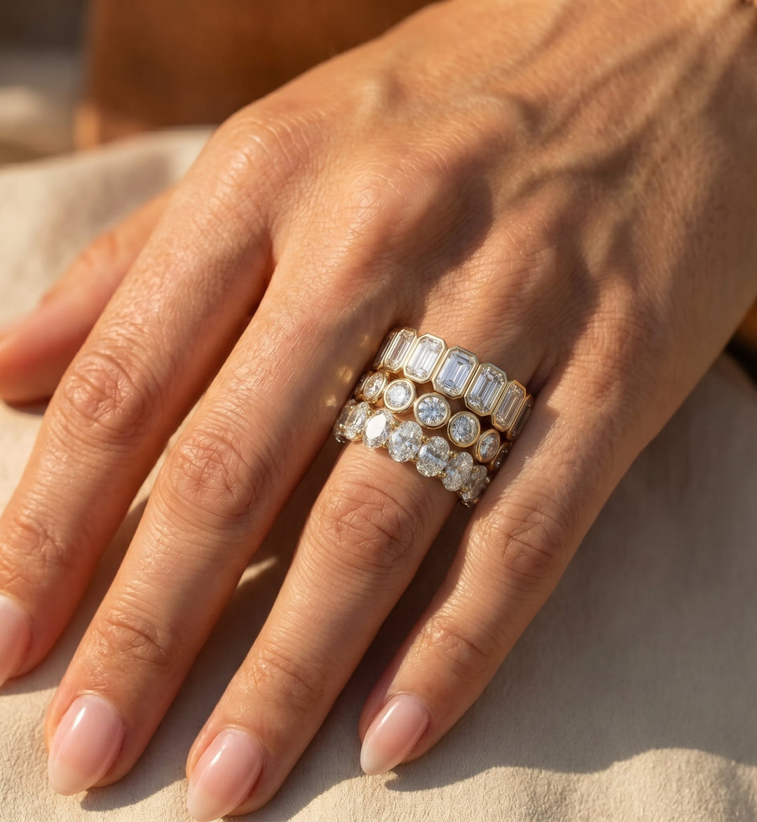 Close-up of a hand adorned with three eternity rings, the top one featuring bezel set emerald cut lab-grown diamonds, followed by a round cut ring, and a full oval eternity  in a classic prong setting - The Jewel Concierge