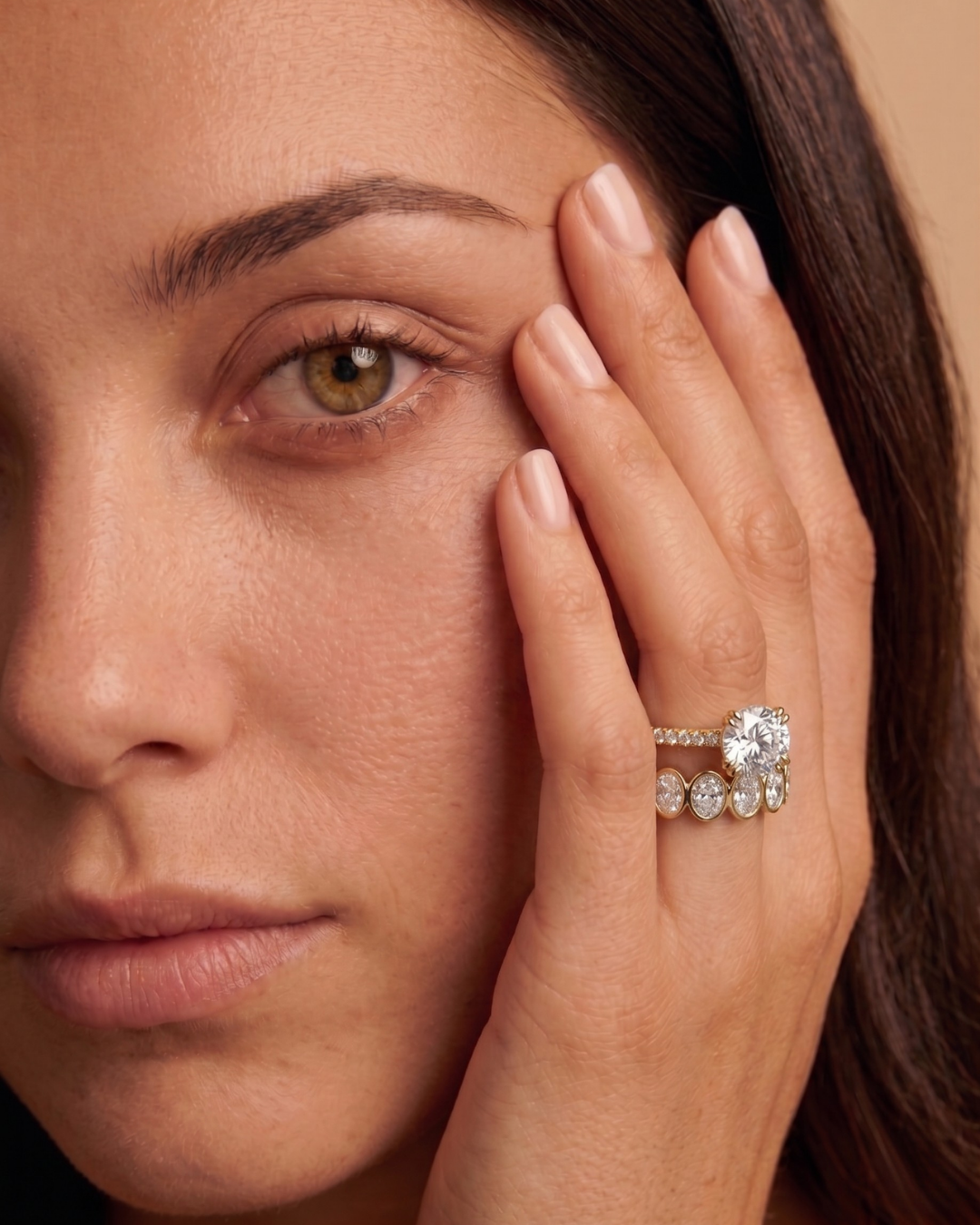 Close-up of a woman, her hand gracefully resting on her face while her fingers adourned with a double claw round cut lab grown diamond engagement ring and an oval bezel set eternity band in 18K yellow gold.