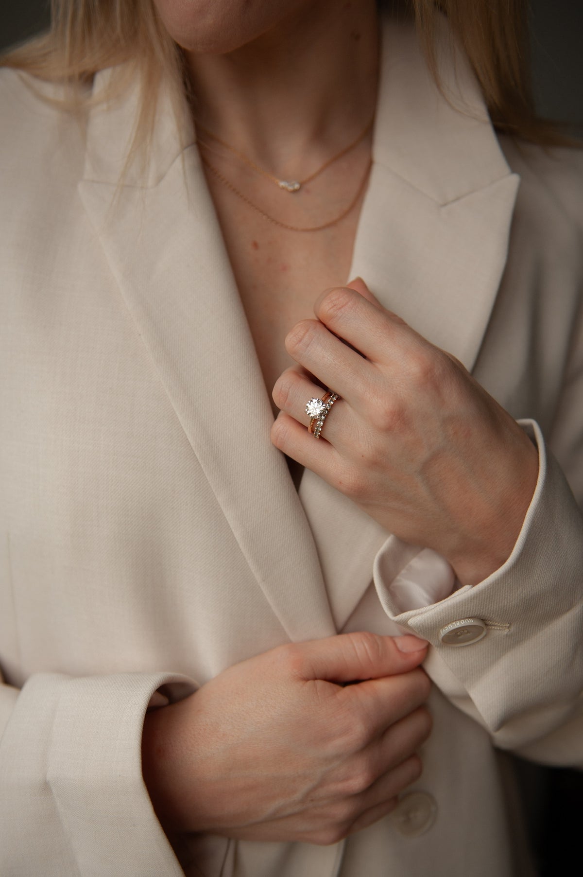 Lifestyle close up image of a model wearing a beige blazer, showcasing a round cut lab-grown diamond engagement ring in 18k yellow gold, stacked with an 18k white gold pave wedding band | The Jewel Concierge
