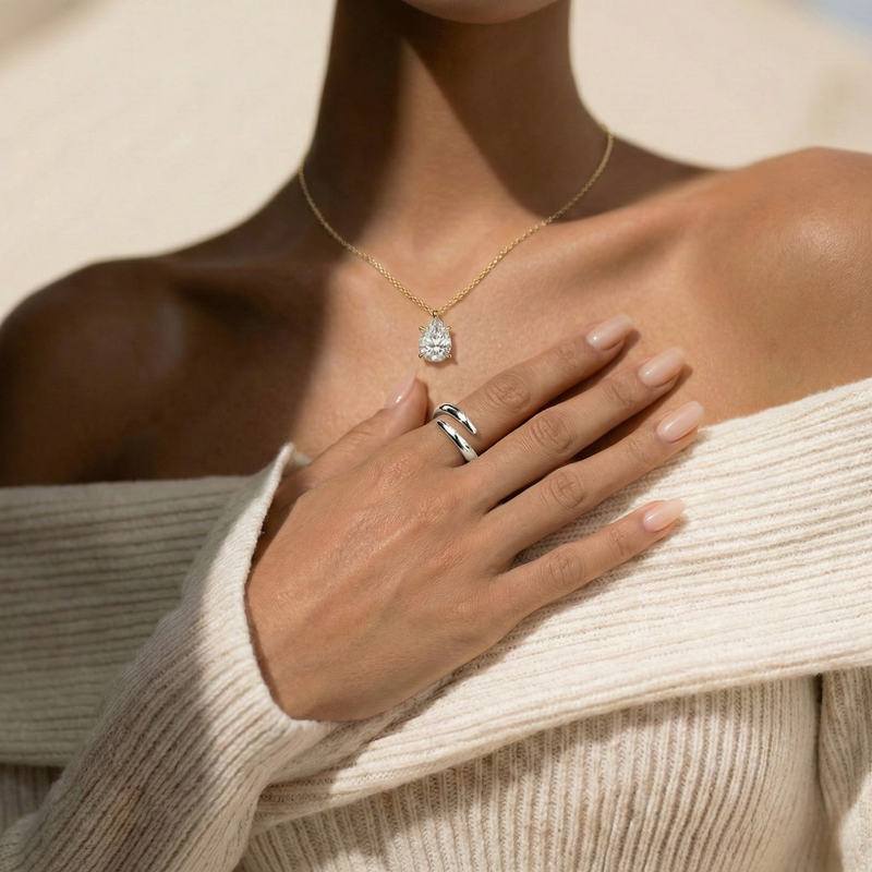 Close-up of a person wearing a pear cut diamond pendant and an open wrap around gold ring, with a neutral background.