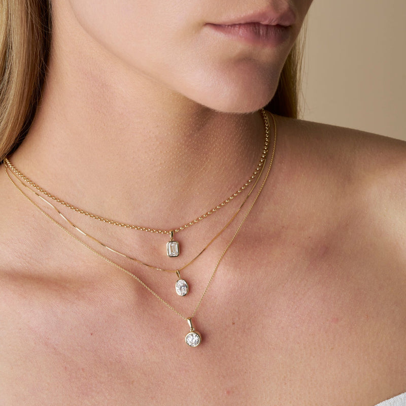Close-up of a model wearing three gold necklaces featuring The Odette Pendant in different diamond shapes, paired with a white ribbed off-the-shoulder top against a neutral beige background - The Jewel Concierge