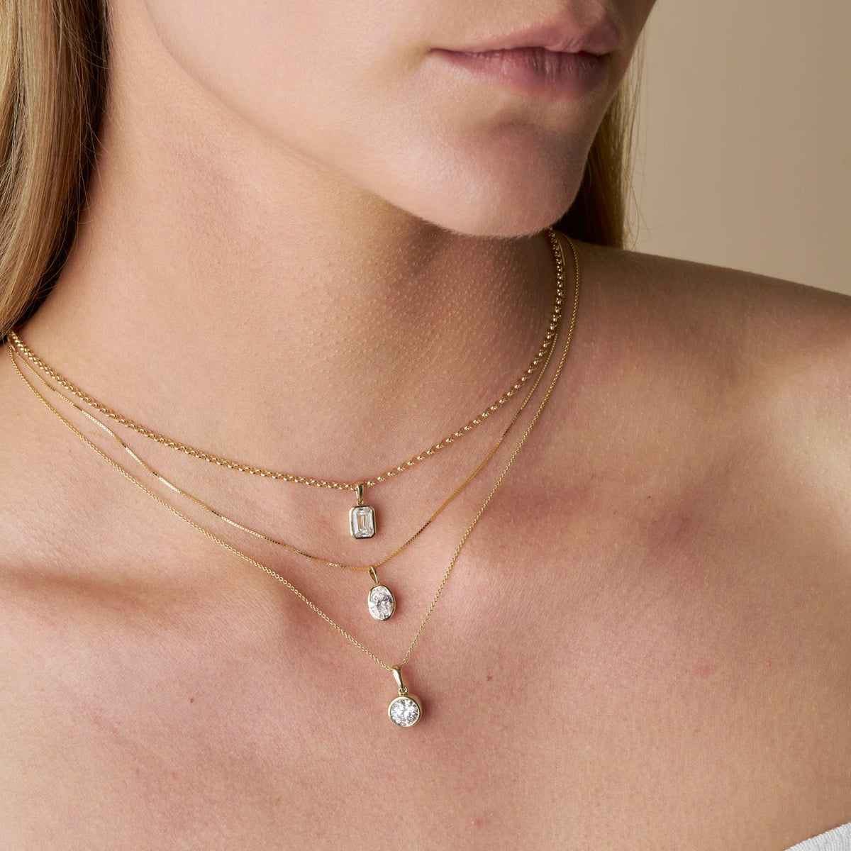 Close-up of a model wearing three gold necklaces featuring The Odette Pendant in different diamond shapes, paired with a white ribbed off-the-shoulder top against a neutral beige background - The Jewel Concierge