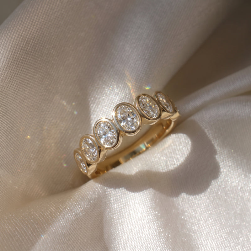 The Sara Ring — Oval Bezel Set Half Lab-Grown Diamond Eternity Band in Yellow Gold on a luxurious silk fabric| The Jewel Concierge