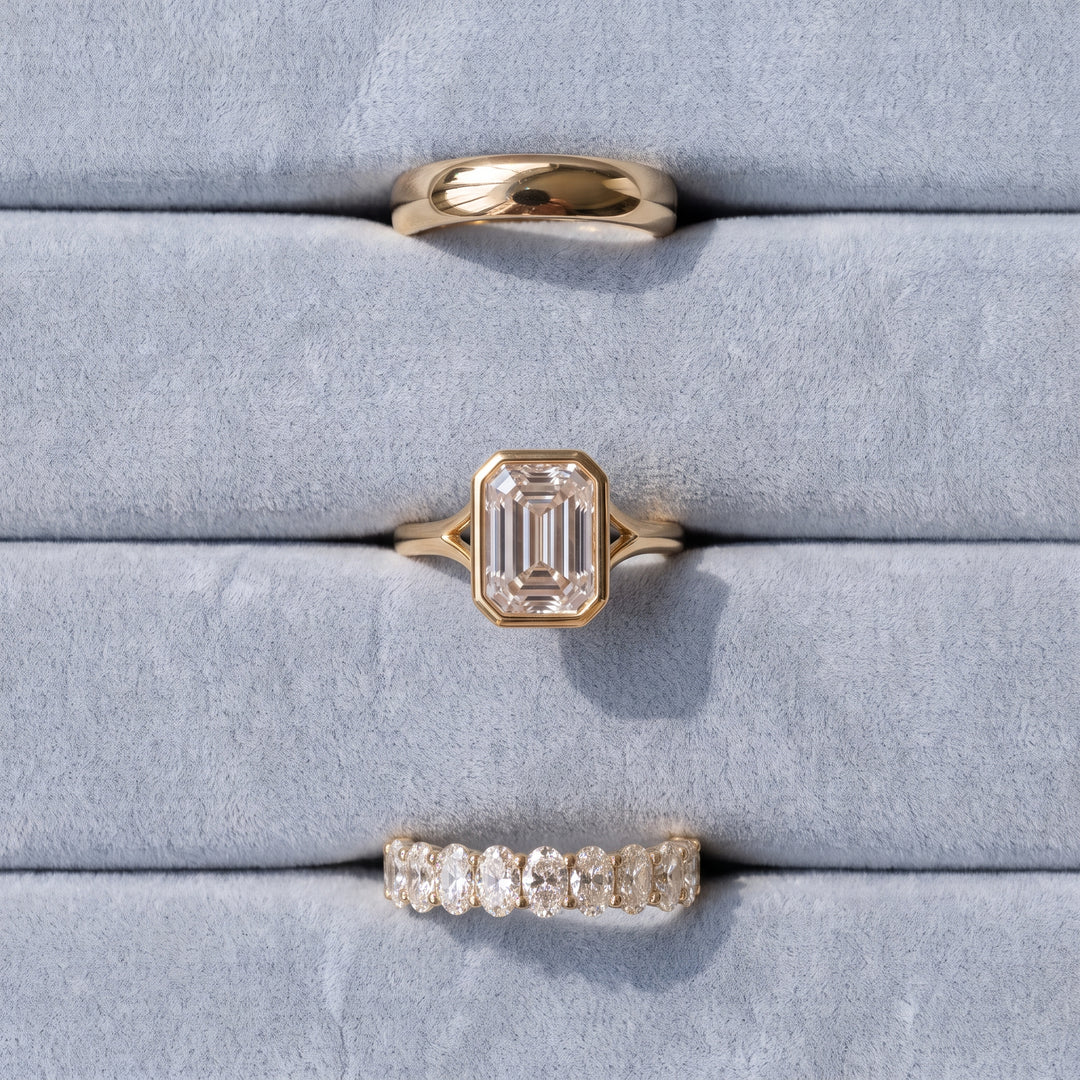 Three rings elegantly placed on a light blue cushion, featuring The Cloud Ring in yellow gold, The Gabriella Ring with an Emerald Solitaire in yellow gold, and The Michelle Oval Eternity Ring in yellow gold | The Jewel Concierge