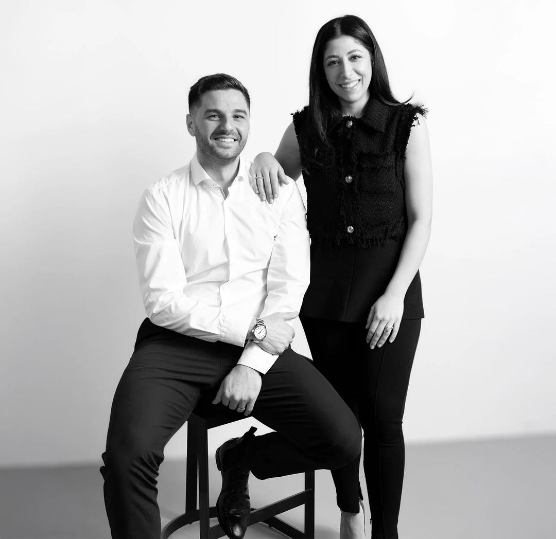 Black and white photo of a man sitting on a stool and a woman standing next to him against a plain background