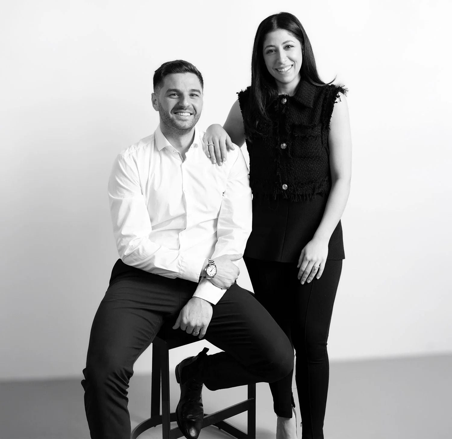 Black and white photo of a man sitting on a stool and a woman standing next to him against a plain background