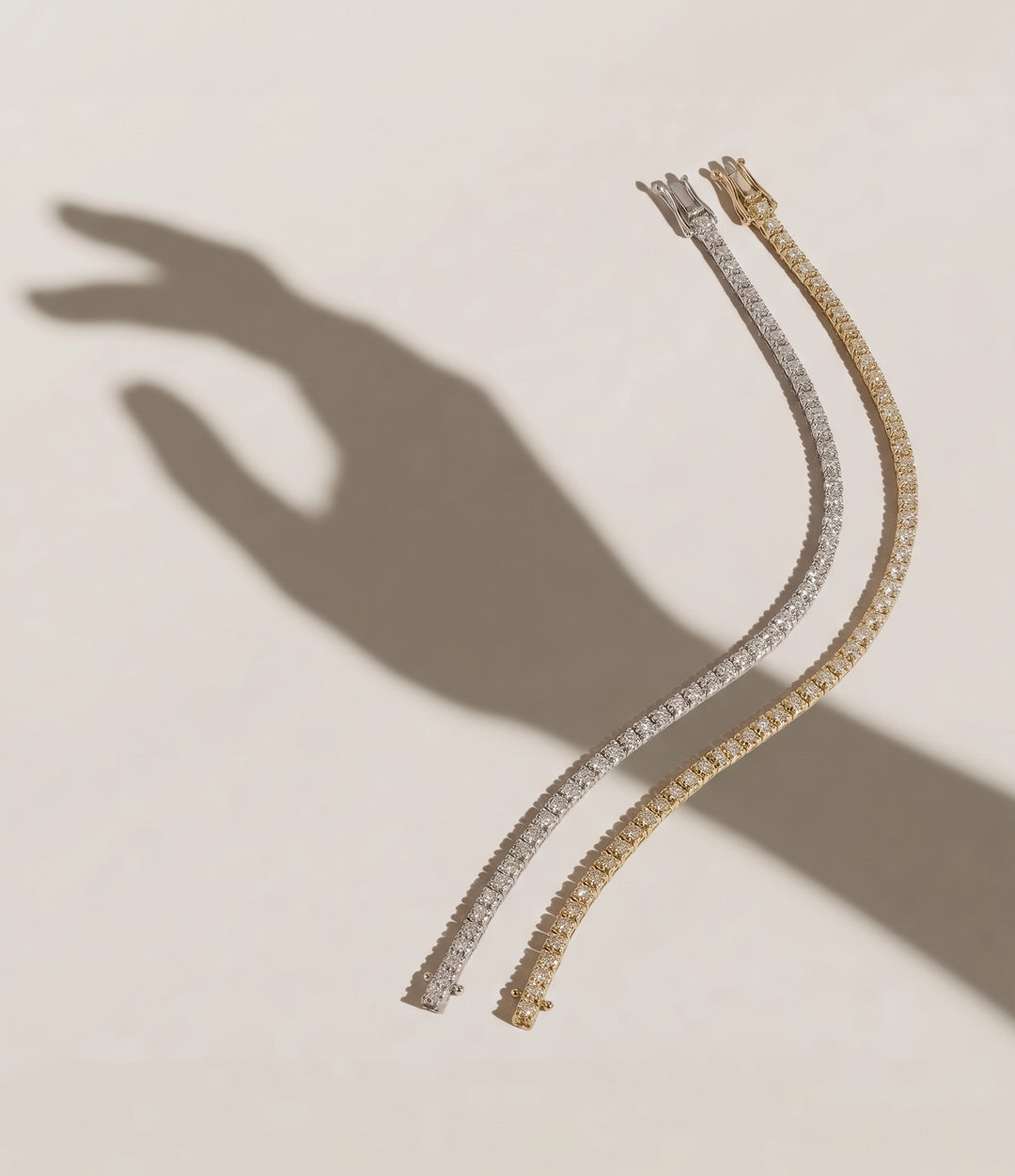 Two Signature tennis bracelets, one in yellow gold and one in white gold, displayed on a warm beige background with a hand shadow cast to the left | The Jewel Concierge