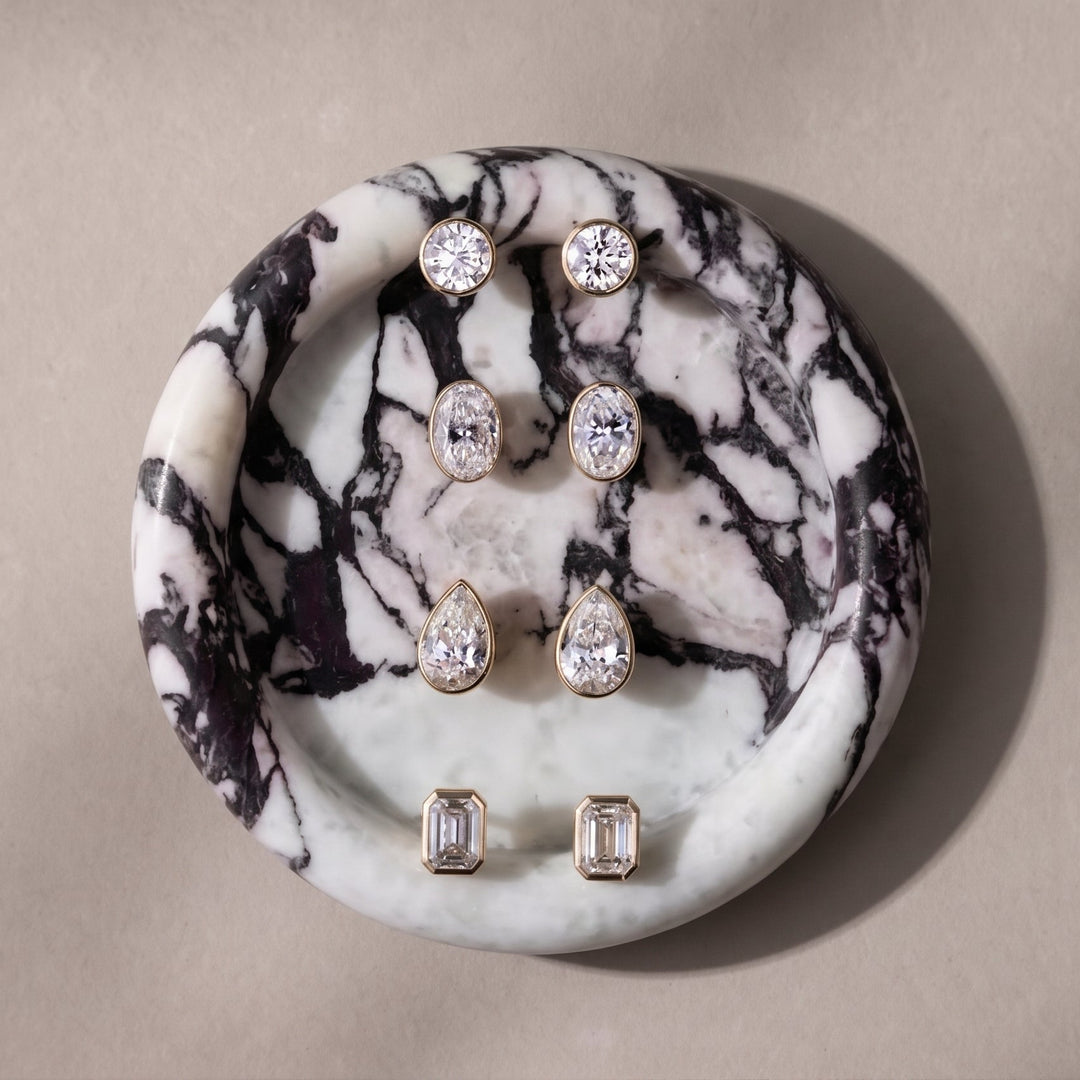 Four pairs of The Aurelia Stud Earrings displayed on a round calacatta viola marble tray against a warm beige background. The earrings feature lab-grown diamonds in a bezel setting of yellow gold, including round, pear, and emerald cuts.