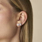 A close-up side profile of a woman's ear wearing The Aurelia Bezel-set studs in emerald and round cut lab-grown diamonds, against a soft beige background - The Jewel Concierge