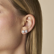 A close-up side profile of a woman's ear wearing The Aurelia Bezel-set studs in emerald and round cut lab-grown diamonds, against a soft beige background - The Jewel Concierge