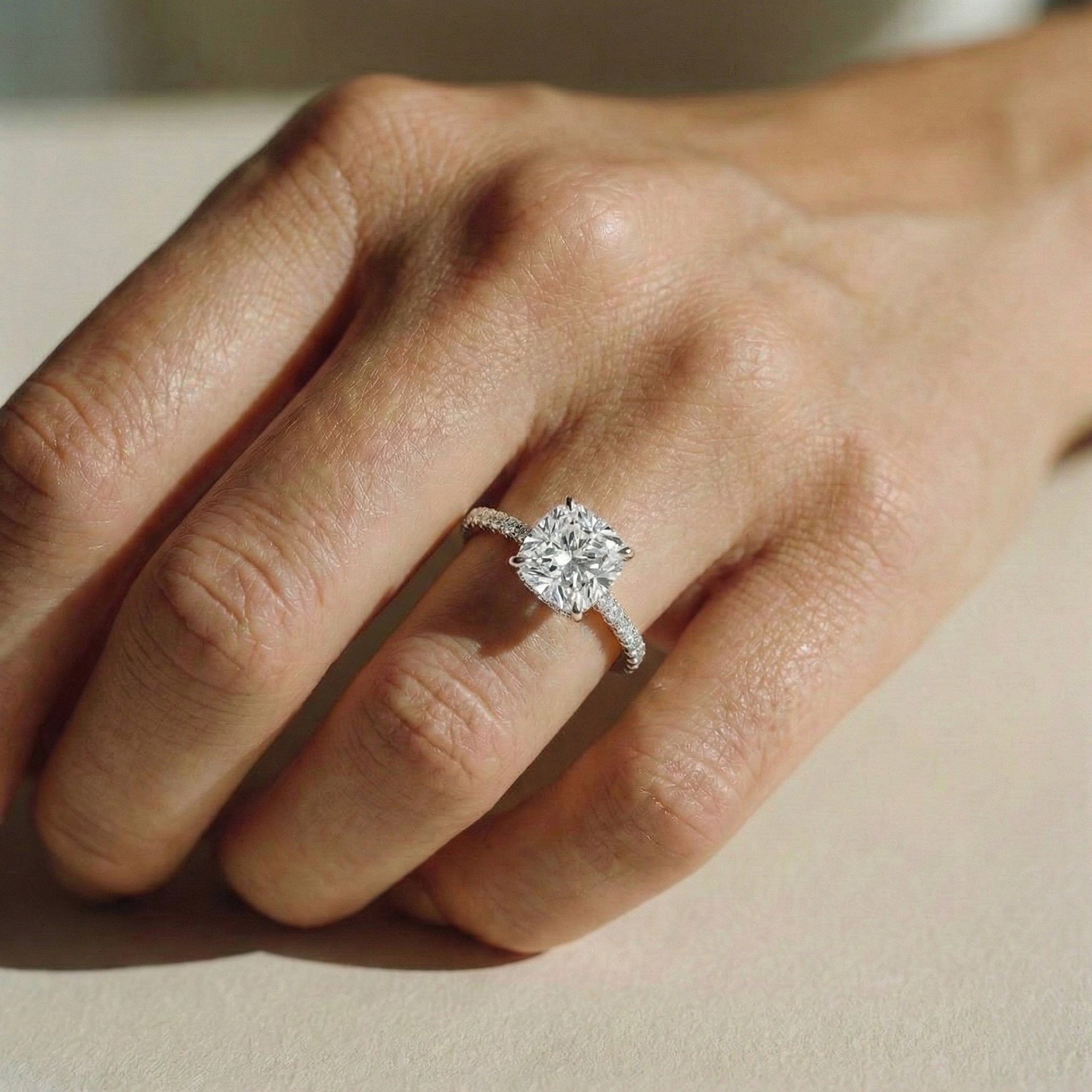 editorial photo of a model's hand resting on a table, wearing The Astra Ring - Cushion Solitaire with pave band in 18K White gold - The Jewel Concierge
