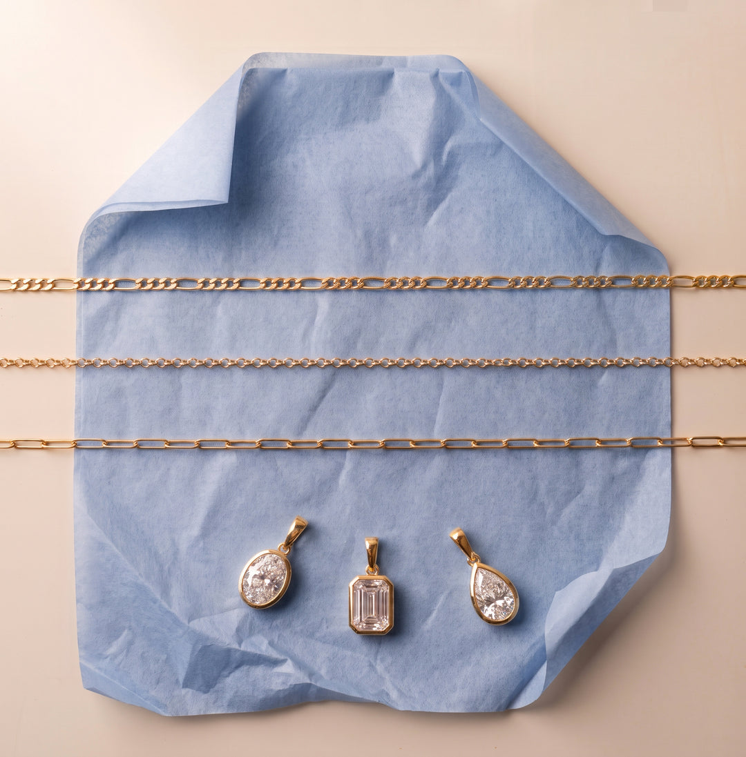 Assortment of fine gold chains and bezel set pendants, elegantly laid on a slate blue parchment paper against a light beige background | The Jewel Concierge