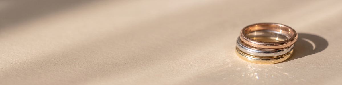 The Emily Ring - A stack of three gold half rounded wedding rings in yellow, white, and rose gold, resting on a textured beige surface with soft shadows | The Jewel Concierge
